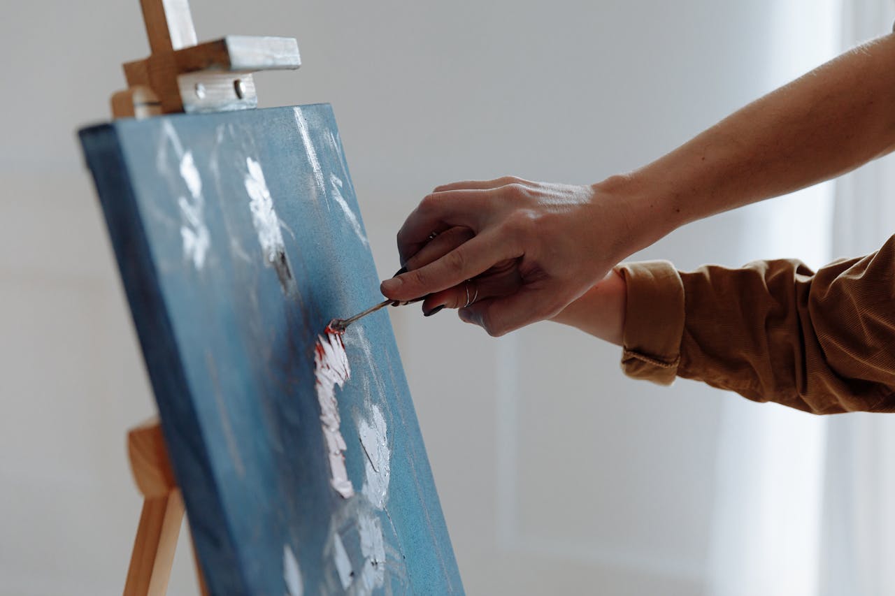 Close-up of a person painting with a spatula on a blue canvas indoors.
